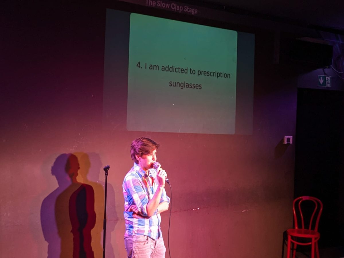 A young comedian performs on stage with a microphone in front of an audience.