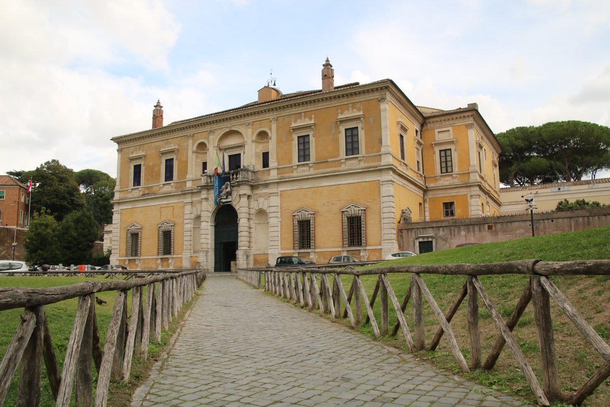 The image shows a yellow museum building with a pathway leading up to it and a wooden fence.