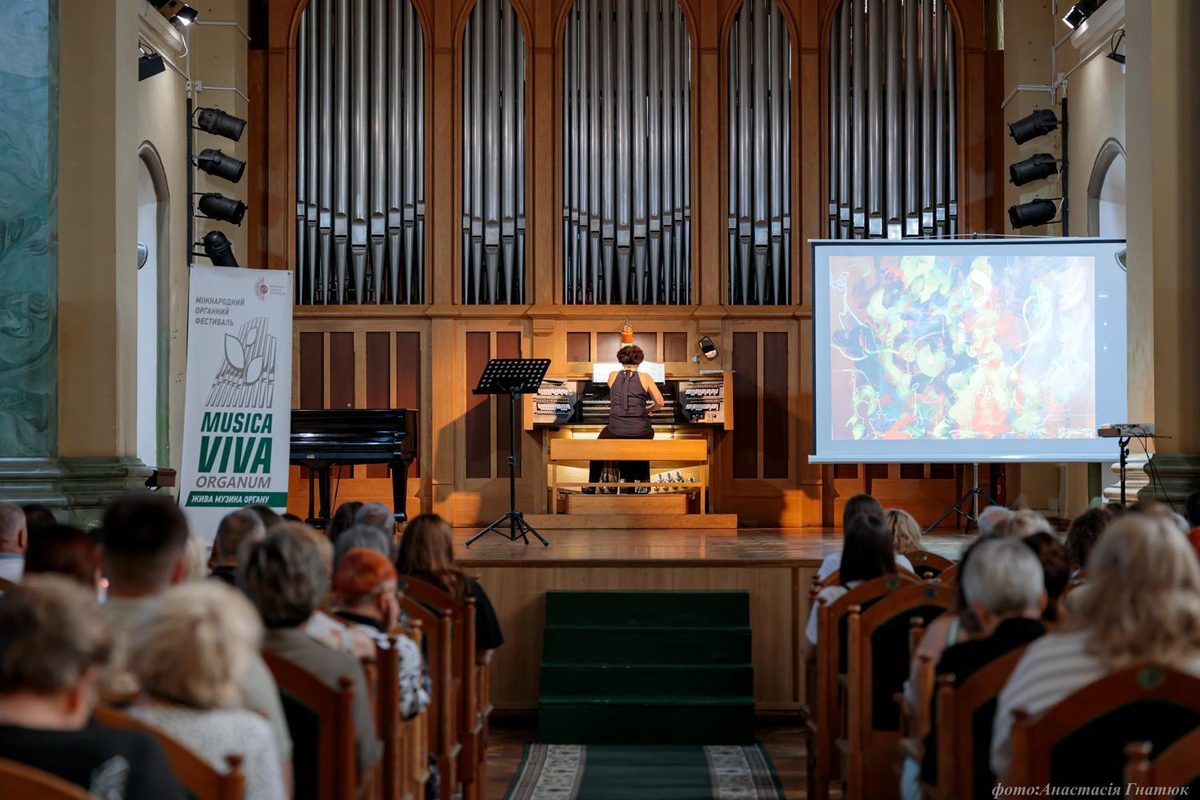 In the concert hall, an audience listens to a musician performing on the organ.