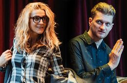 Zwei Personen stehen auf der Bühne, eine Frau mit lockigem Haar in einer Brille und ein Mann in einem Hemd.