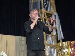 Un uomo parla sul palco con un microfono, e una struttura di legno è visibile dietro di lui.