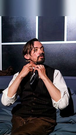 Un hombre con traje clásico se está atando una corbata frente a un fondo oscuro.