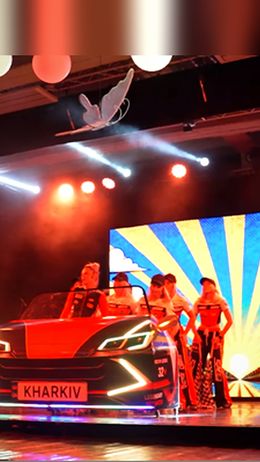 Ein rotes Auto mit der Aufschrift 'KHARKIV' steht auf der Bühne, umgeben von Menschen in Kostümen.